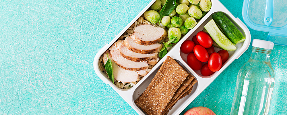 offene Lunchbox mit Hähnchenbrust, Gemüse, Knäckebrot, daneben Apfel und Wasserflasche