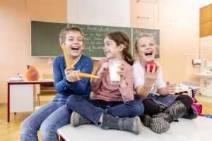 Grundschulkinder sitzen lachend mit Karotten, Milch und Apfel in der Hand auf einem Schultisch