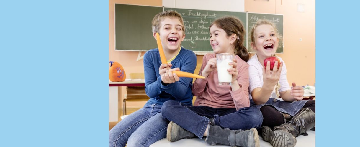 zwei Grundschülerinnen und ein Grundschüler sitzen fröhlich im Klassenzimmer auf einem Tisch und halten Karotten, einen Apfel und ein Milchglas in der Hand